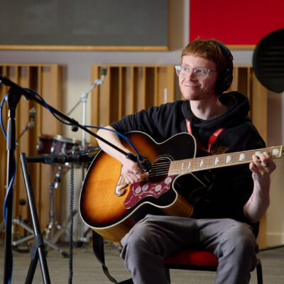 Student sitting in music production studio, using computer and equipment. They are surrounded by a keyboard, microphone, various tech equipment and instruments