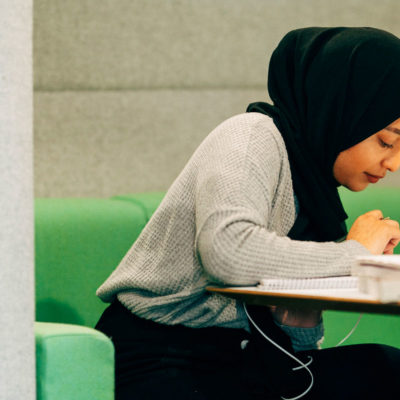 Group of 3 students sitting at a table in a classroom, talking. They have laptops and one is using their hands to gesture while talking.