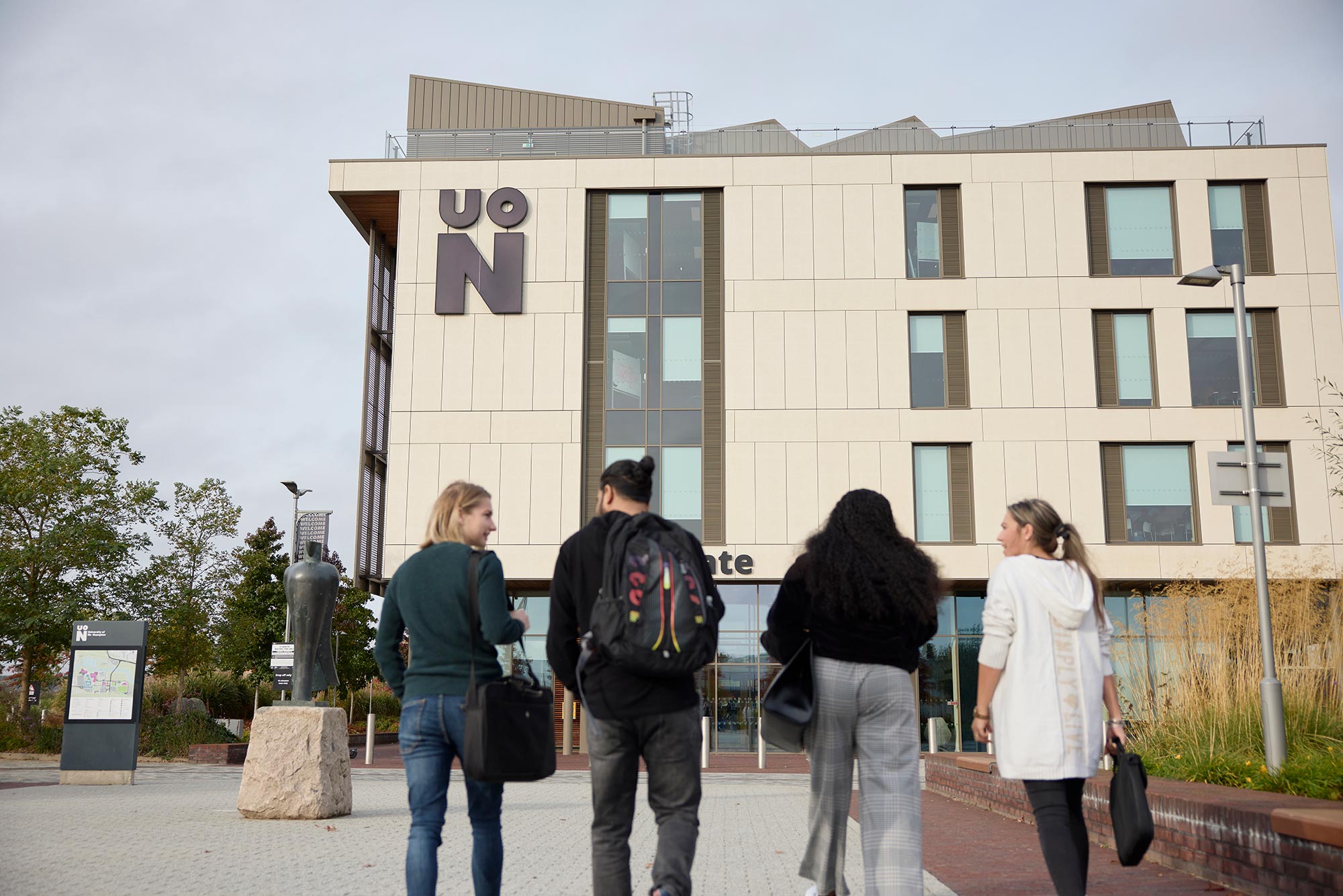 The backs of four students, who are walking away from the camera and towards the Senate building. The UON logo is on the building, as well as a campus map and status outside.