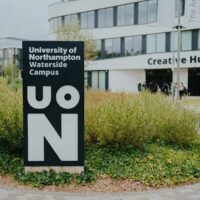 Big sign with 'University of Northampton: Waterside campus' written on it, over UON. The sign is in front of green foliage, which is in front of the Creative Hub building.