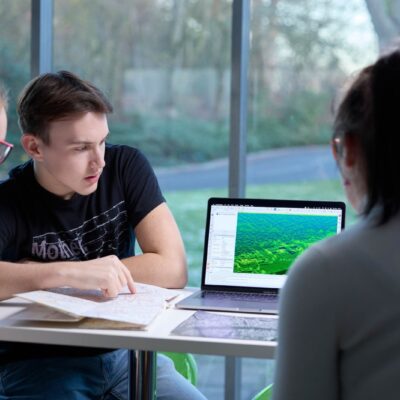 Five students are outside on a field trip. All the students are wearing hi-vis tabards over casual clothes and are looking down at notebooks, phones and clipboards. In the background are trees and blue sky