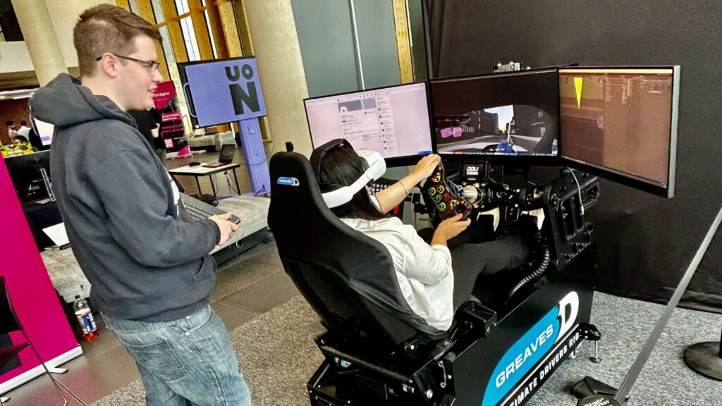 Games Programming Student Russell Waite stands over a driving rig as he guides a visitor through his driving test simulator.