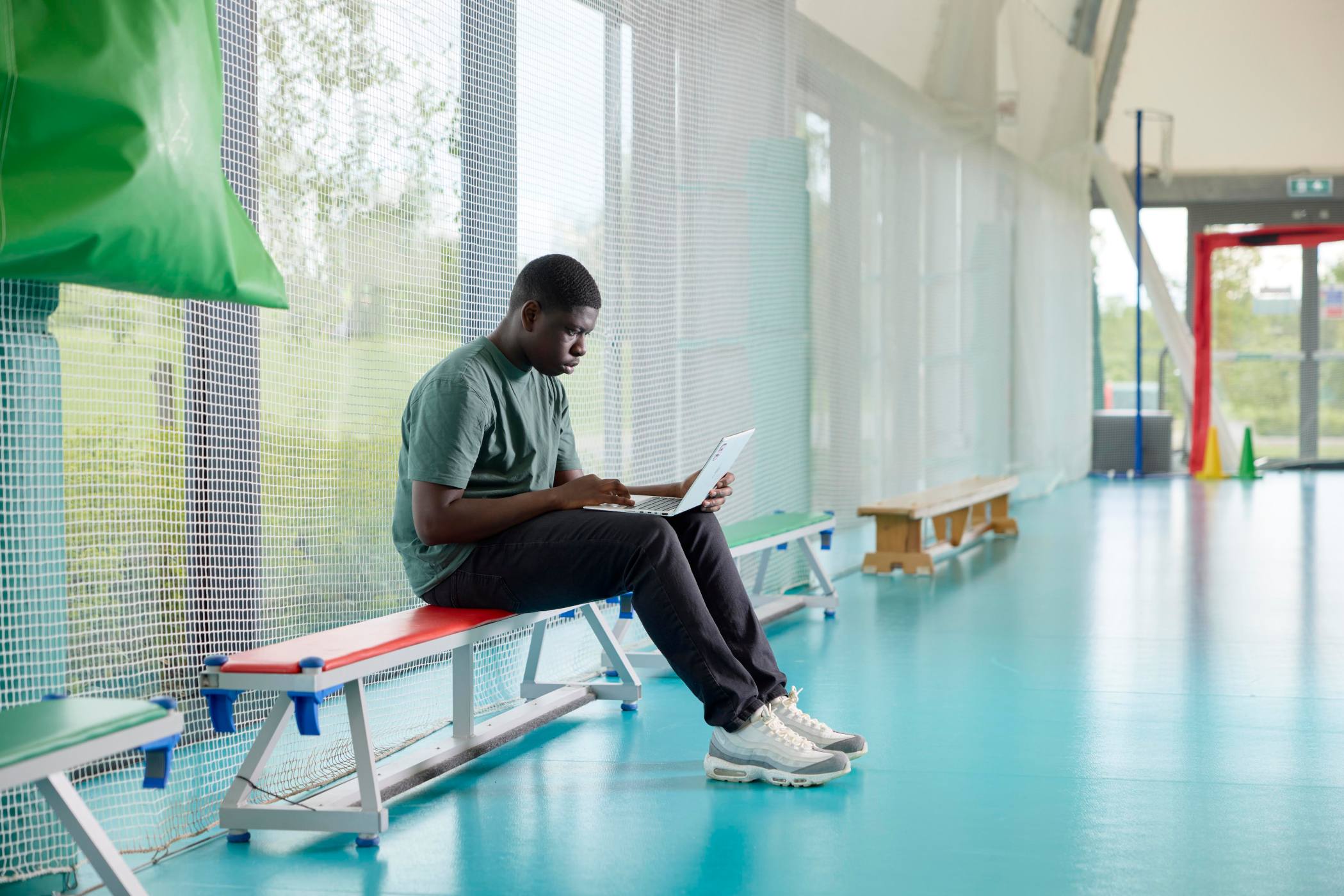 Student sitting on bench in sports hall looking at laptop