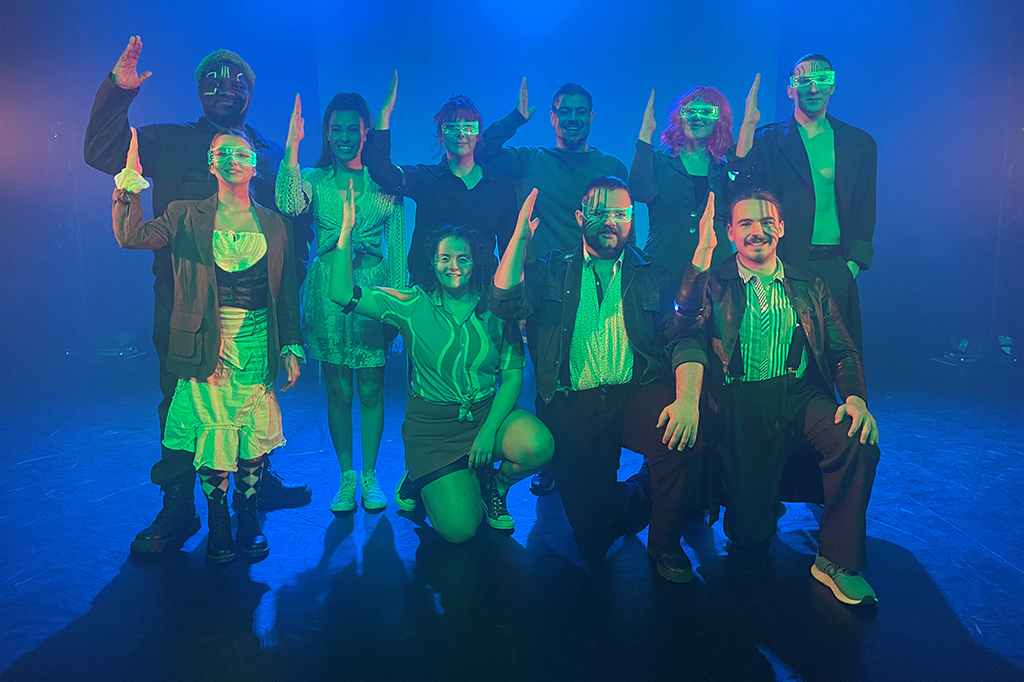 A group of 11 students stand on a stage posing with their right arms raised. They are lit with a green spotlight and the stage background is lit in blue light