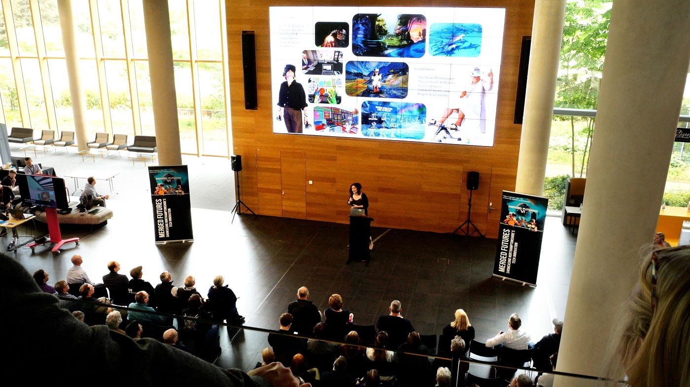 A speaker stands at a podium addressing an audience on the ground floor of the Learning Hub during Merged Futures 6 conference. Behind the speaker is a large screen displaying colourful images.