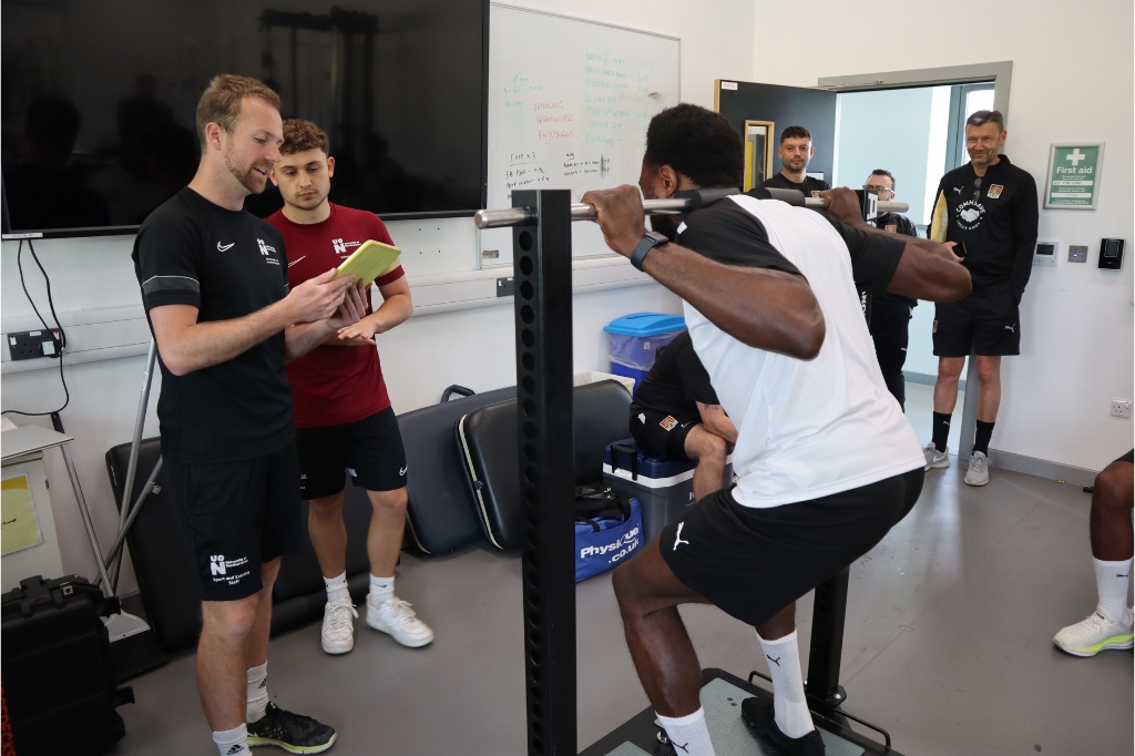 UON staff stand in front of NTFC player as he lifts weight.