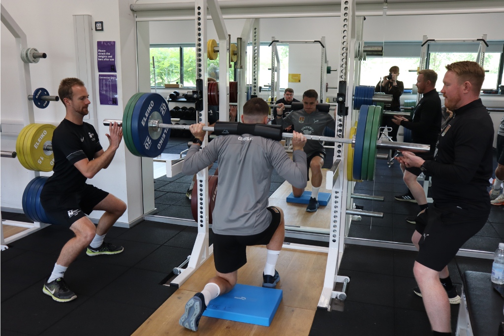 UON staff stand in front of NTFC player as he lifts weight.