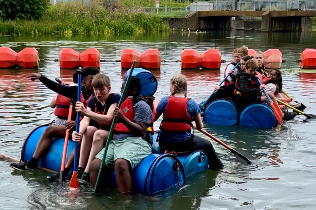Young people from The Ferrers School and Huxlow Academy astride plastic barrels roped together as a raft on the River Nene at Northampton Active whitewater centre.