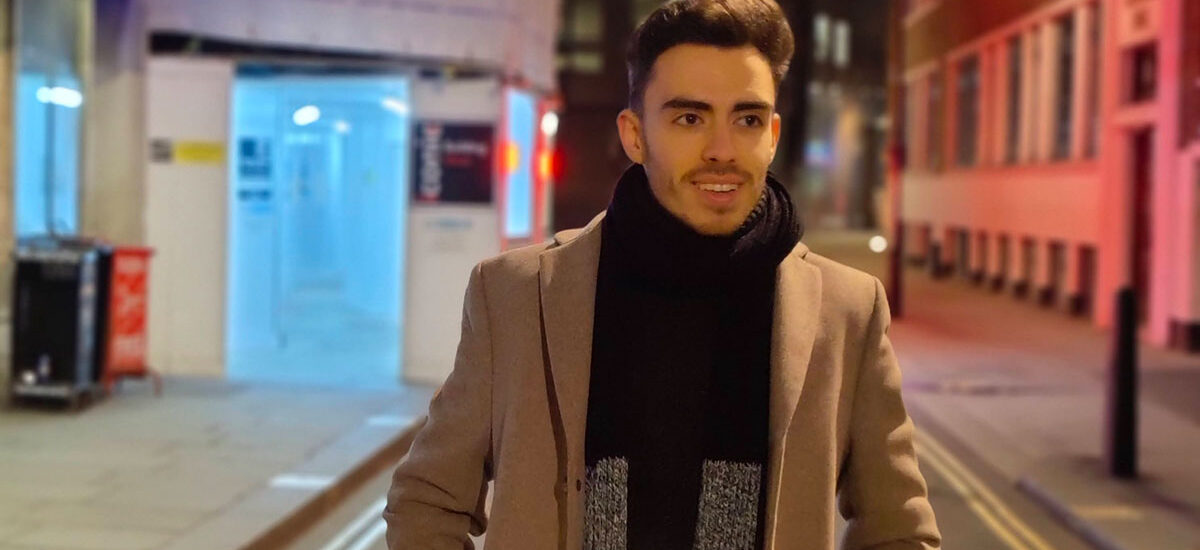 Paulo Figueiredo wearing a beige coat and a black scarf stands on a city street at night, with colorful lights and buildings in the background.