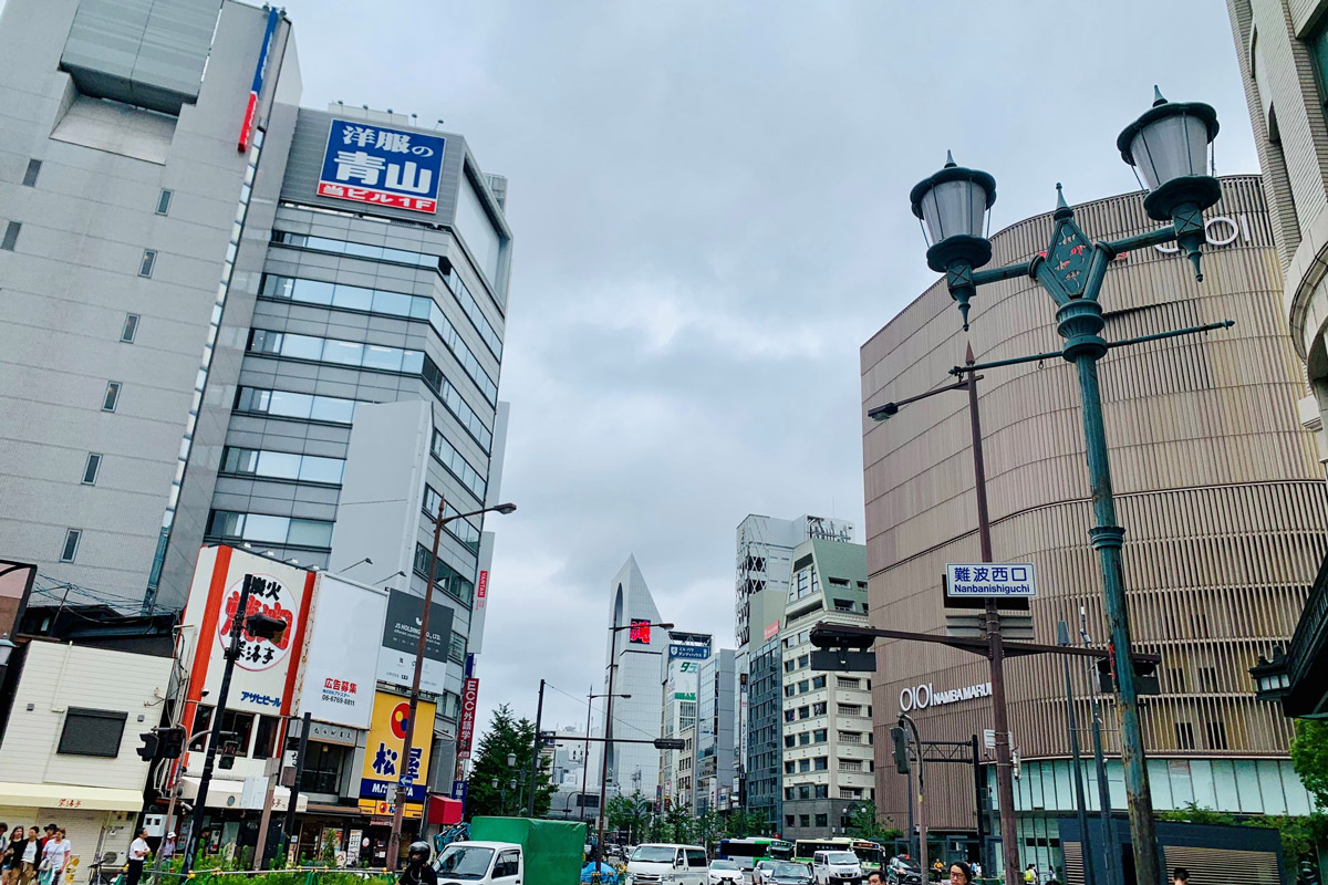 A bustling city street in Japan with tall buildings on both sides, various signs in Japanese, and a modern tower in the background. Street lamps and traffic lights are visible.