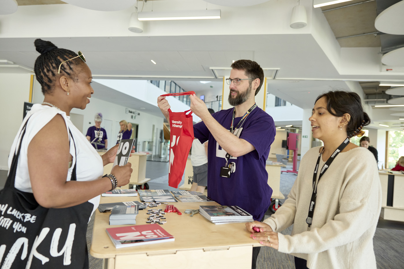 Three people stand around a registration table with pamphlets and materials. One person in a purple shirt holds up a red bag, showing it to the other two. They are in a spacious, well-lit indoor area.