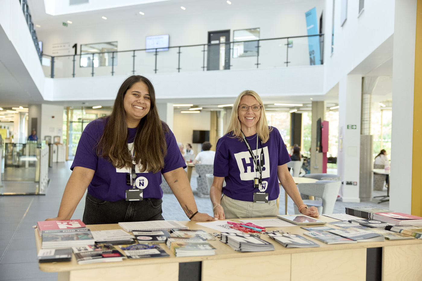 Two people wearing purple UON 'Hi' shirts stand behind a registration desk with brochures in the creative hub entrance, which is well-lit and spacious.