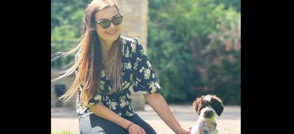 Brooke Savage wears sunglasses and kneels on some grass, smiling beside a small black and white dog wearing a patterned harness. A blurred garden is in the background.