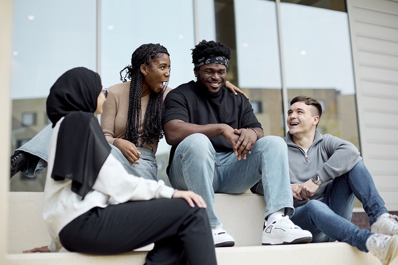 Four students sit on steps outside the Learning Hub, smiling and talking. One woman in a hijab faces the group, while two others engage in conversation with a man in a black shirt in the centre.