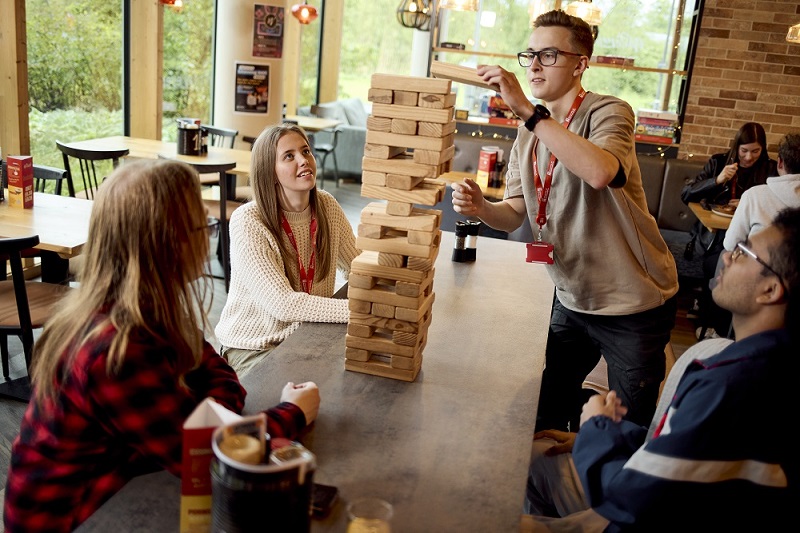 A group of students playing a game of giant Jenga at a table in a casual indoor setting. A student wearing glasses and a light-coloured t-shirt is standing to place a Jenga piece on the top of a stack, while his seated friends watch.