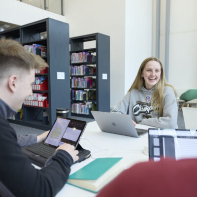 Group of 3 students sitting at a table in a classroom, talking. They have laptops and one is using their hands to gesture while talking.