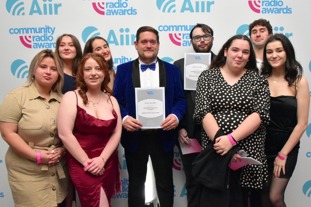 Eight students and NLive Station Manager smile at camera and hold award.