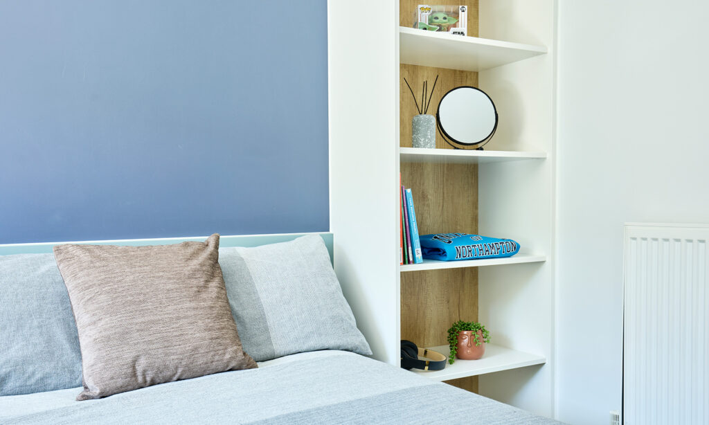 A neatly made bed with gray and beige pillows is next to a white shelf holding books, a small plant, a mirror, and decorative items in a bedroom with a blue accent wall.