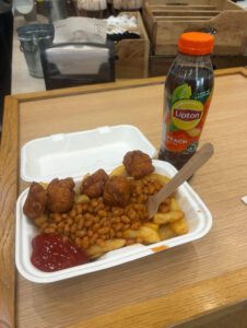 Chicken bites, chips ,and beans in a white food container on a table, with a Lipton peach iced tea next to it.