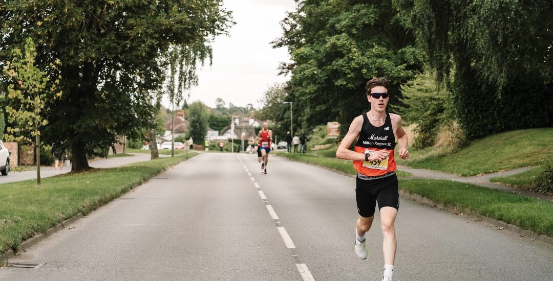 Sport Rehabilitation and Conditioning student, Joshua Edwards running in a marathon on a road. He is ahead, wearing sunglasses and a number taking part in the sport.