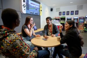 Four students sit at a table and talk in the Waterside restuarant games room. In the background are arcade machines against a back wall, and a large screen showing motor racing.