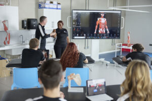 A group of students sit in a classroom observing a digital anatomical model on a large screen. At the front of the class, an academic holds the arm of a student as part of a demonstration.