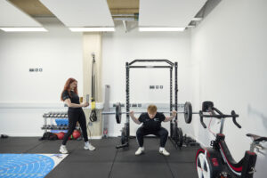 A person squats with a barbell across their shoulders in UON’s SIRC while another person stands nearby, observing. Exercise equipment including a bike, dumbbell rack, and squat rack are visible in the background.