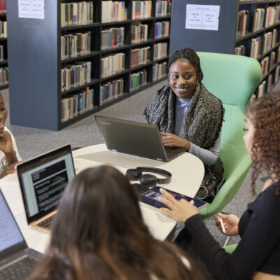 A student sitting at a table, writing in a notepad. They are looking up and directly at the camera, smiling. In the background, other students are seen at desks in a blur.