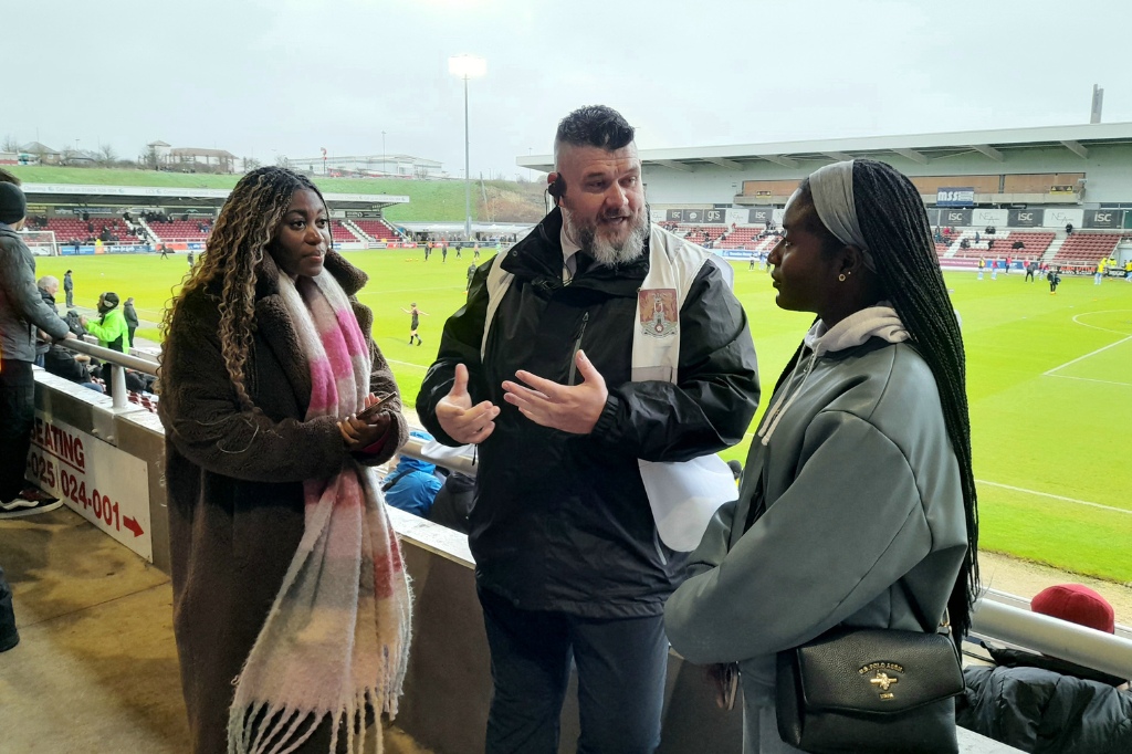 Students stand and chat with security personnel in front of NTFC football pitch in the background.