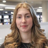 Grace Harrison, standing in between two bookshelves looking at the camera and smiling.