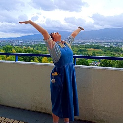 Student Samantha O'Neill is wearing a blue dress and stands on a balcony with arms outstretched, overlooking a scenic landscape in Japan with hills and cloudy sky.