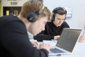 Two people wearing headphones are focused on their laptops at a table.