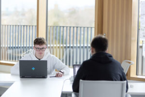 Two people sit across from each other at a table, both working on laptops, in a bright room with large windows.