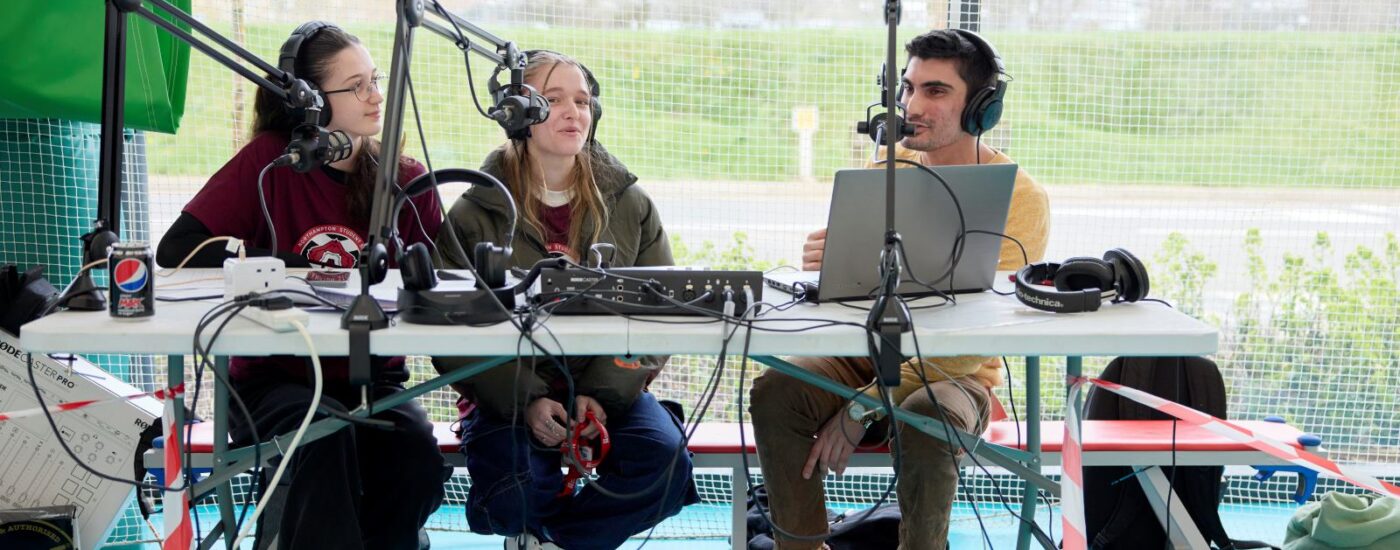 Three students sitting at a table with microphones and headphones, engaged in a discussion or podcast recording. Audio equipment, laptop, and a drink are placed on the table.